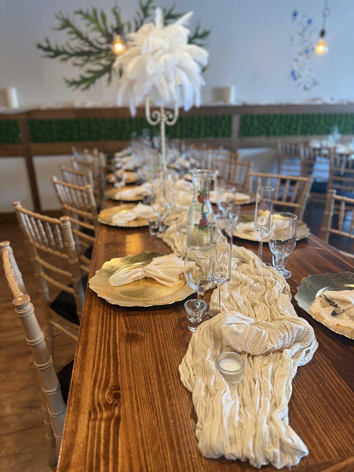 Long table set with gold chargers and a greenery wall